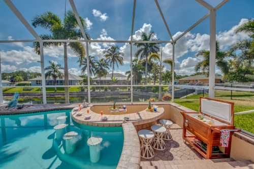 A sunny pool area with a bar, stools, and palm trees, enclosed by a screen for a tropical vibe.