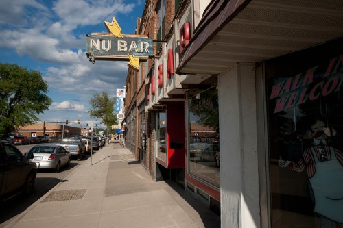 A street view featuring the "Nu Bar" sign, parked cars, and storefronts under a partly cloudy sky.