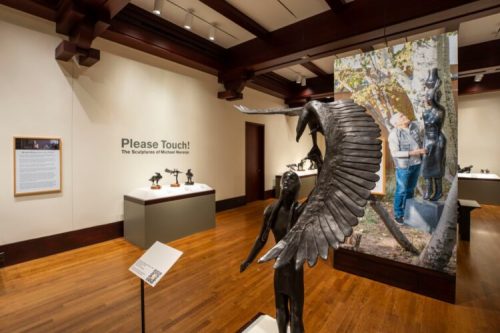 A gallery featuring sculptures by Michael Kienzer, with a "Please Touch!" sign and a visitor interacting with a piece.