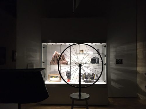 A dimly lit museum display featuring a large wheel in front of various historical artifacts in glass cases.