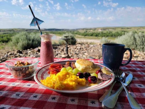A breakfast spread on a picnic table with eggs, fruit, a smoothie, and coffee, set against a scenic landscape.