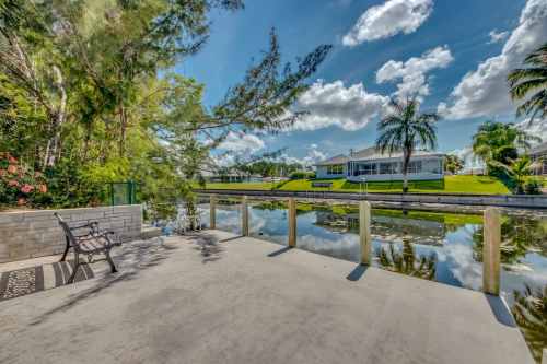 A serene waterfront view with a concrete patio, palm trees, and reflections on the calm water under a blue sky.