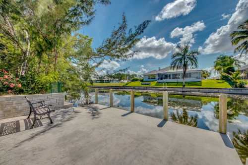 A serene waterfront view with a concrete patio, palm trees, and reflections on the calm water under a blue sky.