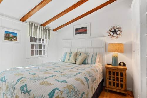 Cozy bedroom with a white bedspread featuring fish, wooden beams, a lamp, and nautical decor on the walls.