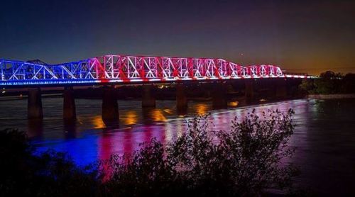A bridge illuminated in red, white, and blue lights at night, reflecting in the water below.