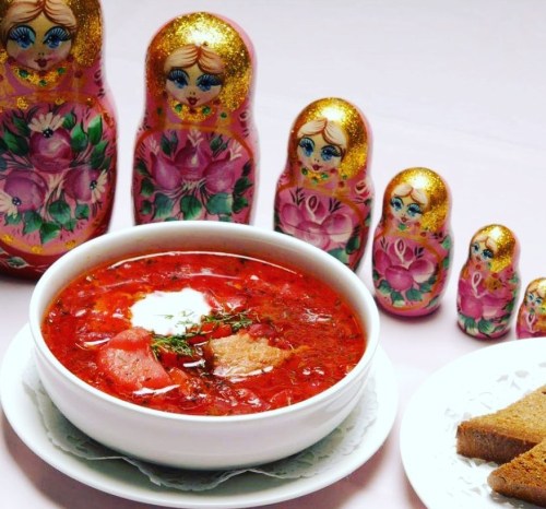 A bowl of borscht topped with sour cream, surrounded by colorful Russian nesting dolls and slices of bread.