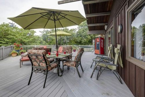 A spacious deck with a table, chairs, and umbrellas, surrounded by greenery and featuring a Coca-Cola vending machine.