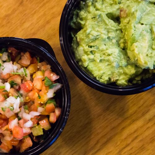 A black bowl of fresh pico de gallo next to a bowl of creamy guacamole on a wooden surface.