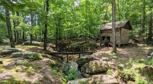 A rustic wooden cabin surrounded by lush green trees and rocks, with a small bridge over a stream nearby.