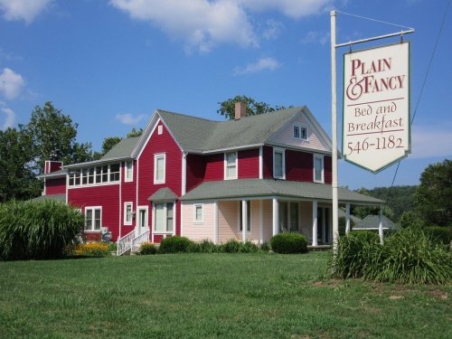 A charming red and pink bed and breakfast with a sign, surrounded by green grass and trees under a blue sky.
