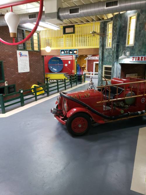A vintage red fire truck inside a colorful, child-friendly museum space with various play areas.