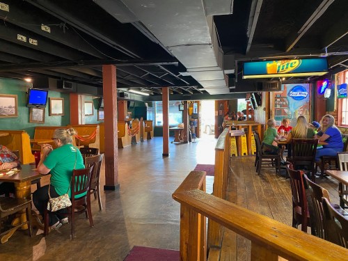 Interior of a casual restaurant with wooden tables, patrons dining, and a bar area in the background. Bright and inviting atmosphere.