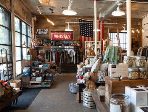A rustic store interior featuring clothing racks, home goods, and decor, with an American flag and whiskey sign.