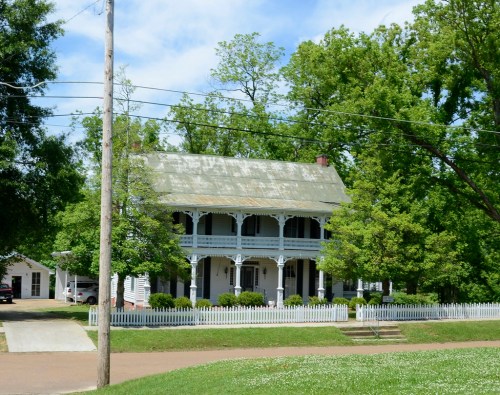 A large, two-story white house with a green roof, surrounded by trees and a white picket fence.