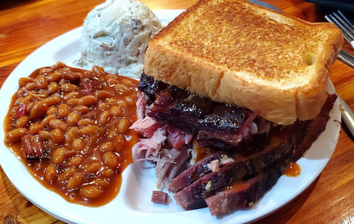 A plate featuring a stacked sandwich with meats, baked beans, and a side of creamy coleslaw.