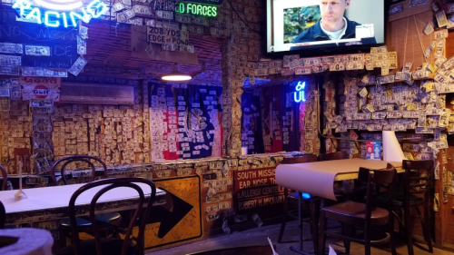 A cozy bar interior filled with dollar bills and memorabilia on the walls, featuring a TV and wooden chairs.