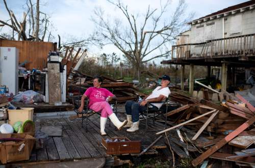 Two people sit on a deck amidst debris and destruction, reflecting the aftermath of a recent storm.
