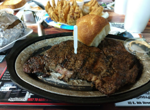 A sizzling steak on a plate with a roll and a side of fries in the background.