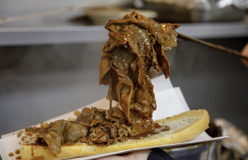 A close-up of a sandwich with piled high, juicy beef slices drizzled in sauce on a crusty bread roll.