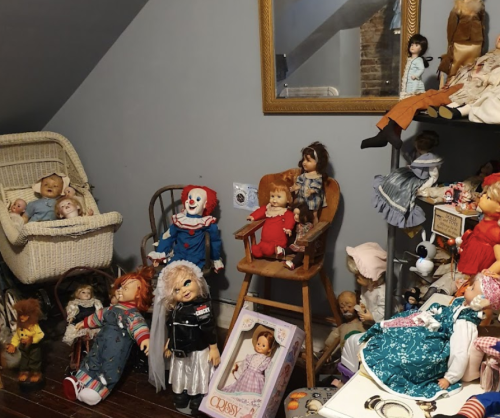 A cluttered room filled with various dolls, including vintage and creepy ones, arranged on chairs and shelves.