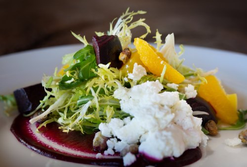 A vibrant salad with greens, beets, orange segments, and crumbled cheese, drizzled with a berry sauce.