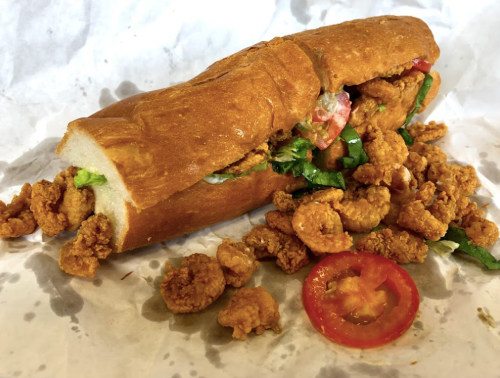 A sandwich filled with fried shrimp, lettuce, and tomato, with extra shrimp spilling out onto the wrapper.