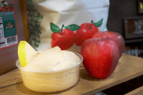 A container of creamy dip next to two red apples and a bag of apple cider mix on a wooden surface.