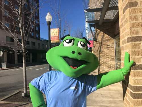 A green frog mascot in a blue shirt poses playfully outside a building on a sunny day.