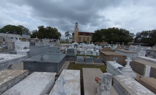 A cemetery with numerous gravestones, featuring a church in the background under a cloudy sky.