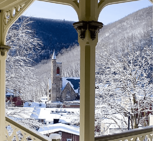 A snowy landscape viewed from a porch, featuring a church steeple and mountains in the background.