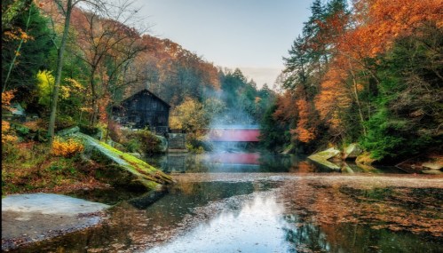 A serene autumn landscape featuring a calm river, colorful trees, and a rustic building with smoke rising in the background.