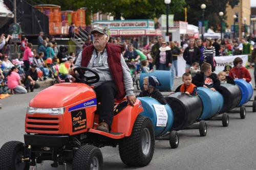A man drives a small tractor pulling children in barrels during a parade, with a crowd watching in the background.