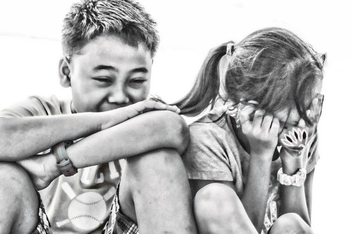 Two children sitting together, one laughing and the other covering their face, in a playful moment. Black and white image.