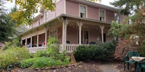 A charming pink house with a porch, surrounded by greenery and autumn leaves, featuring outdoor seating.