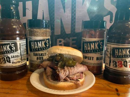 A sandwich with sliced meat and jalapeños, surrounded by Hank's Barbecue sauce and rub bottles on a wooden table.