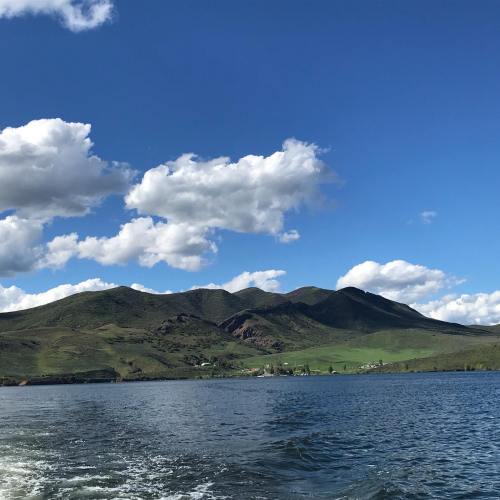 A serene landscape featuring green hills under a blue sky with fluffy white clouds, viewed from a body of water.