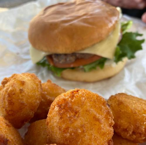 A close-up of golden, crispy tater tots in front of a cheeseburger with lettuce and tomato in the background.