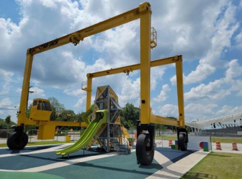 A large yellow crane structure with a slide underneath, set in a playground with blue skies and fluffy clouds.