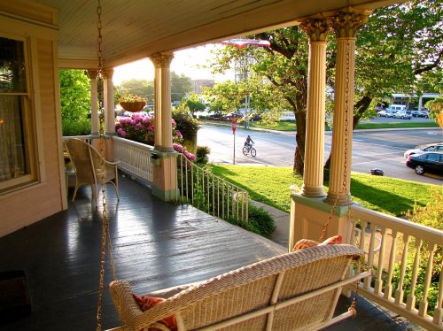 A sunny porch with a swing, overlooking a street where a cyclist rides by, surrounded by greenery and flowers.