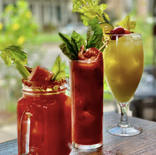 Three colorful cocktails on a wooden table: a Bloody Mary, a tall drink with herbs, and a yellow drink with a cherry.