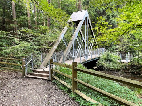 A wooden bridge with a triangular metal frame spans a small creek, surrounded by lush green trees and a wooden fence.