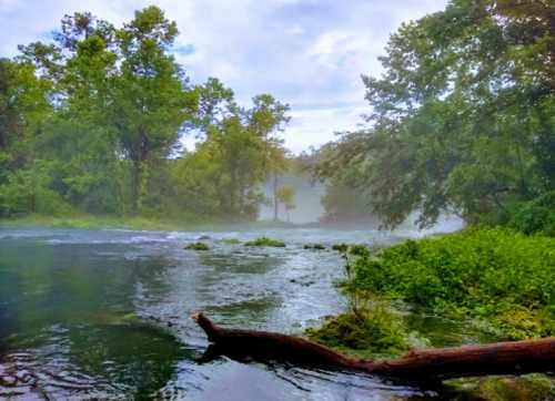 A serene river scene with mist, surrounded by lush greenery and trees under a cloudy sky.