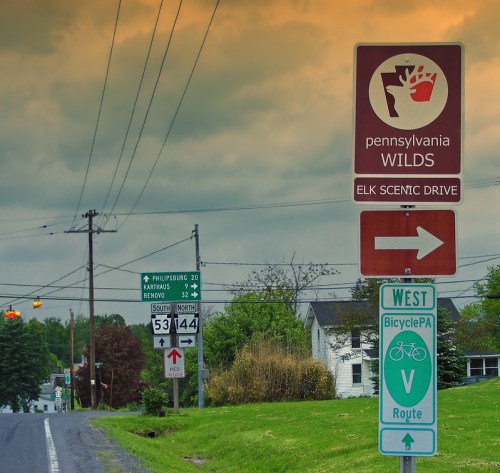 Signpost for Pennsylvania Wilds and Elk Scenic Drive, with directional arrows and nearby road signs under a cloudy sky.