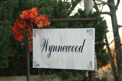 Sign reading "Wynnewood" adorned with autumn leaves and a plaid ribbon, set against greenery.