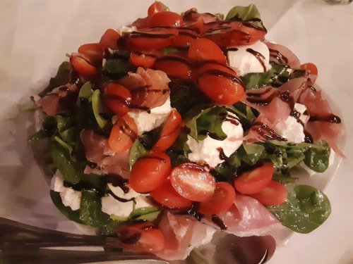 A colorful salad with spinach, cherry tomatoes, prosciutto, and drizzled balsamic glaze on a white plate.