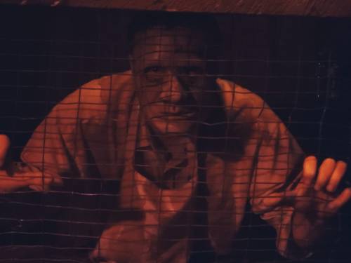 A man peers through a grid, illuminated by dim light, with an intense expression on his face.