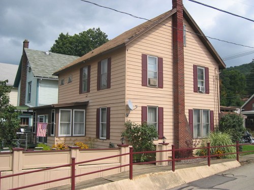 A two-story peach-colored house with maroon shutters, a brick chimney, and a small garden in front.