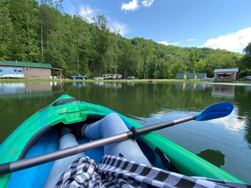 A person in a kayak on a calm lake, surrounded by green trees and a few buildings in the background.