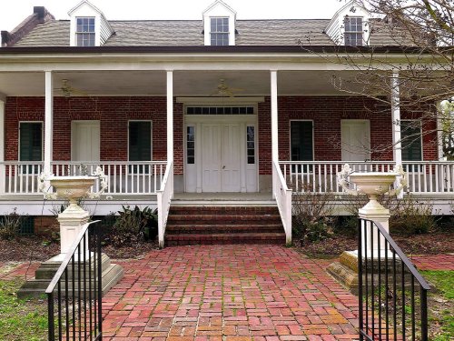 Historic brick house with a large porch, white railings, and two front doors, surrounded by a garden and brick pathway.