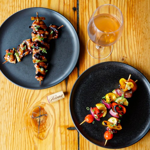 Two plates of skewers: one with grilled meat and vegetables, the other with colorful roasted veggies, alongside a glass of drink.